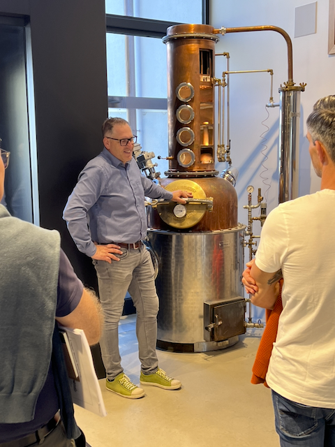 Ein Mann in hellblauem Hemd und grauer Hose steht vor einem grossen, kupferfarbenen Destillierapparat. Er lächelt und gestikuliert mit der Hand in Richtung des Apparats. Zwei Personen stehen im Hintergrund und hören zu. Fokus auf Brenn-Kurs/Destillation.