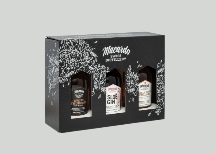 Eine schwarze Geschenkbox mit drei kleinen Flaschen Spirituosen. Die Box ist mit einer weißen Illustration von Pflanzen und Früchten verziert. Die Aufschrift "Macardo Swiss Distillery" ist gut lesbar. Die Flaschen enthalten Whisky, Gin und Eau-de-vie.