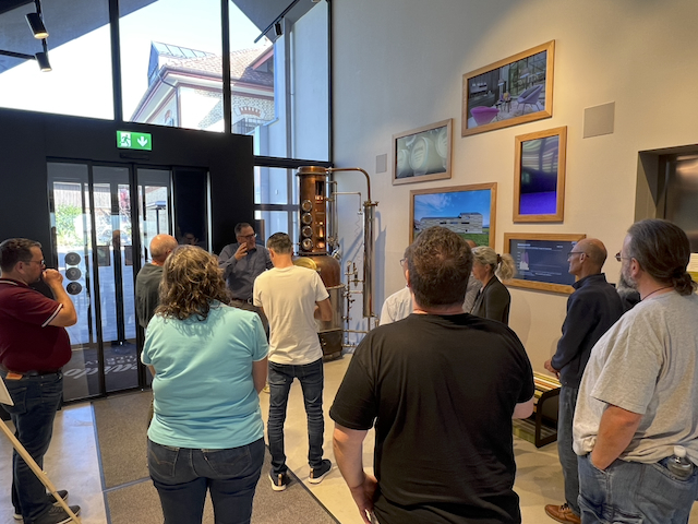 Eine Gruppe steht in einem Raum, vermutlich bei einem Macardo Brennkurs. Im Hintergrund ist eine kupferfarbene Destillationsanlage zu sehen. An der Wand hängen Bilderrahmen mit verschiedenen Motiven. Ein Mann probiert aus einem Glas.