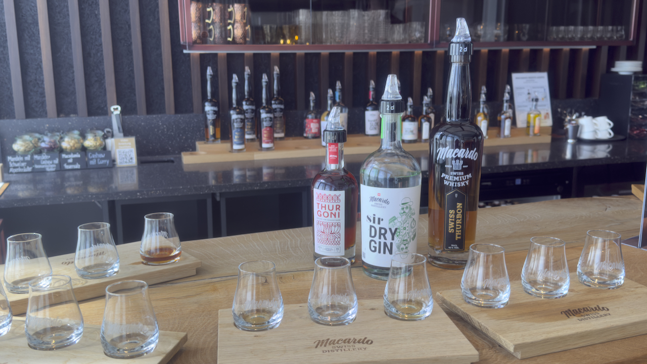 Auf einem Tisch stehen Holztabletts mit kleinen, leeren Gläsern. Dahinter stehen Flaschen mit Spirituosen, darunter Gin und Whisky, von Macardo. Im Hintergrund ist eine Bar mit weiteren Flaschen und Gläsern erkennbar.