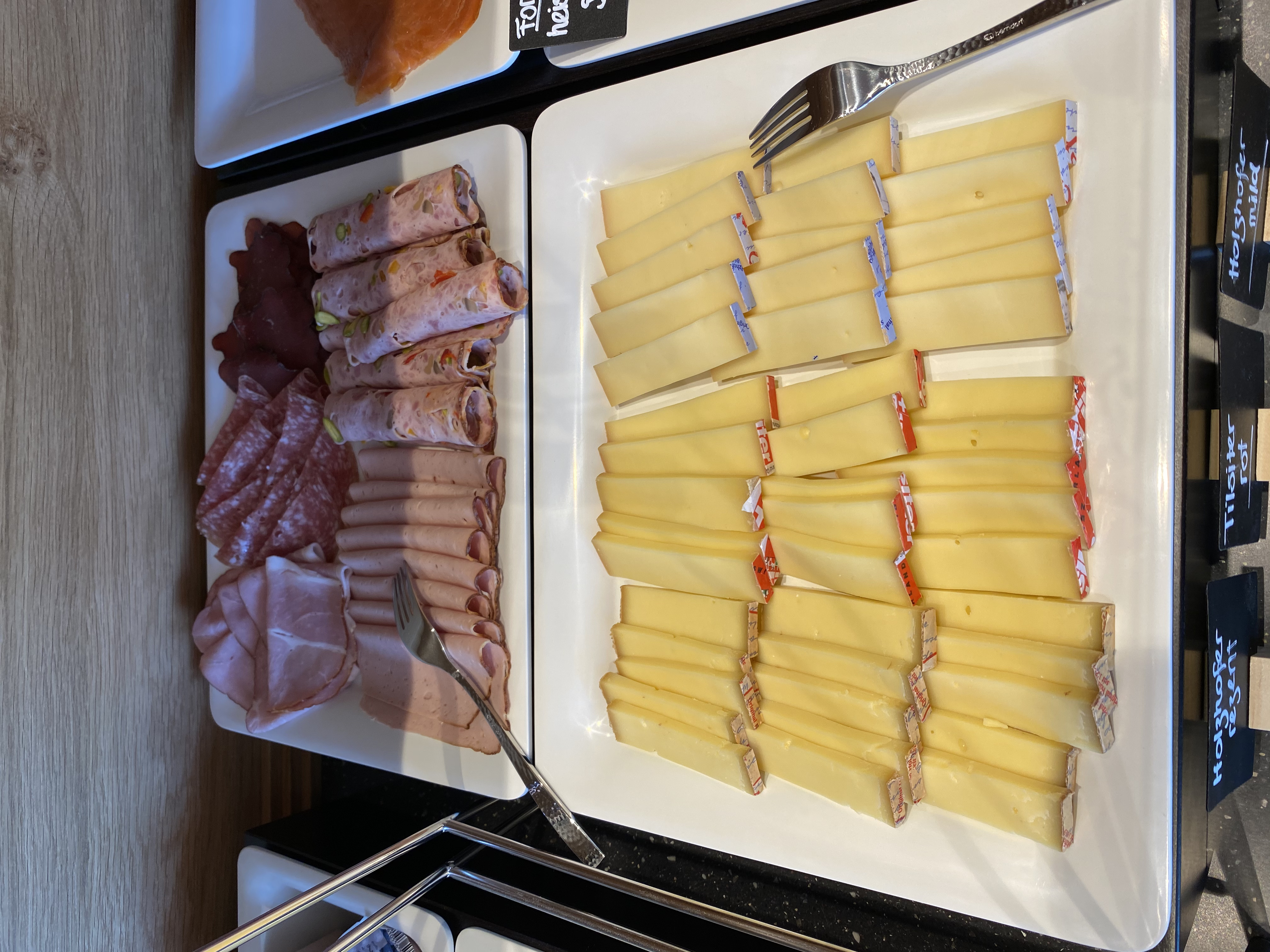 Auf dem Buffet sind Platten mit diversen Käsesorten und Fleischwaren angerichtet. Die Käsesorten sind in Scheiben geschnitten. Es gibt eine Gabel zur Selbstbedienung. Die Aufmachung erinnert an einen reichhaltigen Brunch.