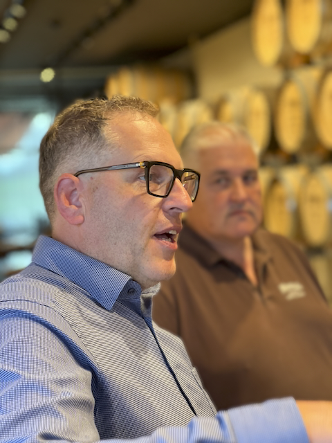 Nahaufnahme zweier Männer, der vordere Mann trägt eine blau-weiss gestreifte Knopfleiste und eine Brille und spricht. Der Mann im Hintergrund trägt ein braunes Poloshirt. Im Hintergrund sind unscharf Holzfässer der Macardo Swiss Distillery zu sehen.