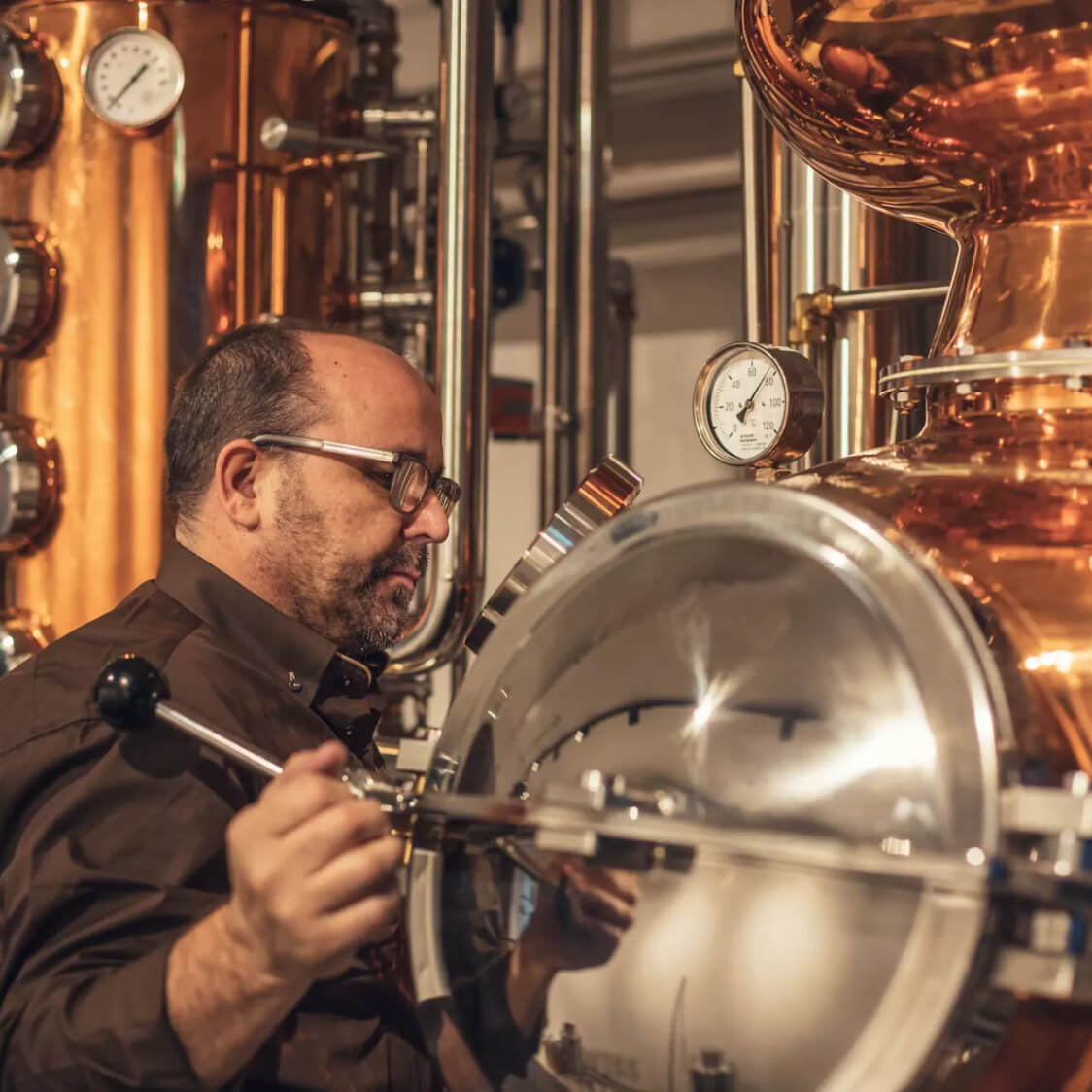 Ein Mann mit Brille, der an einer grossen, silbernen Destillieranlage arbeitet. Im Hintergrund glänzen kupferfarbene Brennkessel und Rohre. Die Szene vermittelt einen Eindruck von Handwerkskunst und Fachwissen im Brennereiwesen bei Macardo.