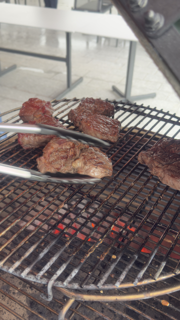 Nahaufnahme von saftigen Steaks auf einem Grillrost über glühenden Kohlen. Eine Grillzange greift nach einem der Stücke. Im Hintergrund sind verschwommen Tische und Stühle erkennbar.
