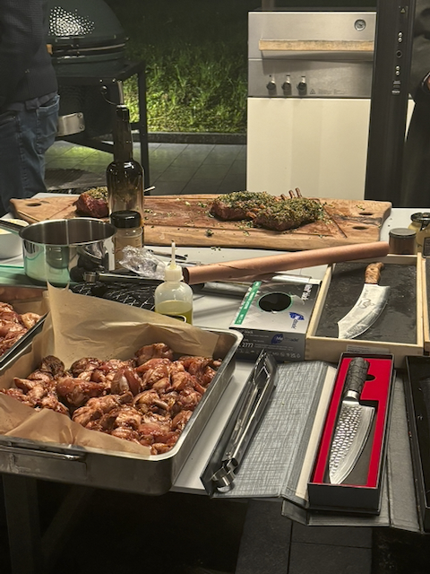 Tisch mit Grillzutaten und Werkzeugen: mariniertes Fleisch in Schale, Schneidebrett mit Lammkoteletts und Kräutern, Ölflasche, Gewürze, Grillzangen und Damastmesser in Schatullen. Im Hintergrund Grill und Außenküche.