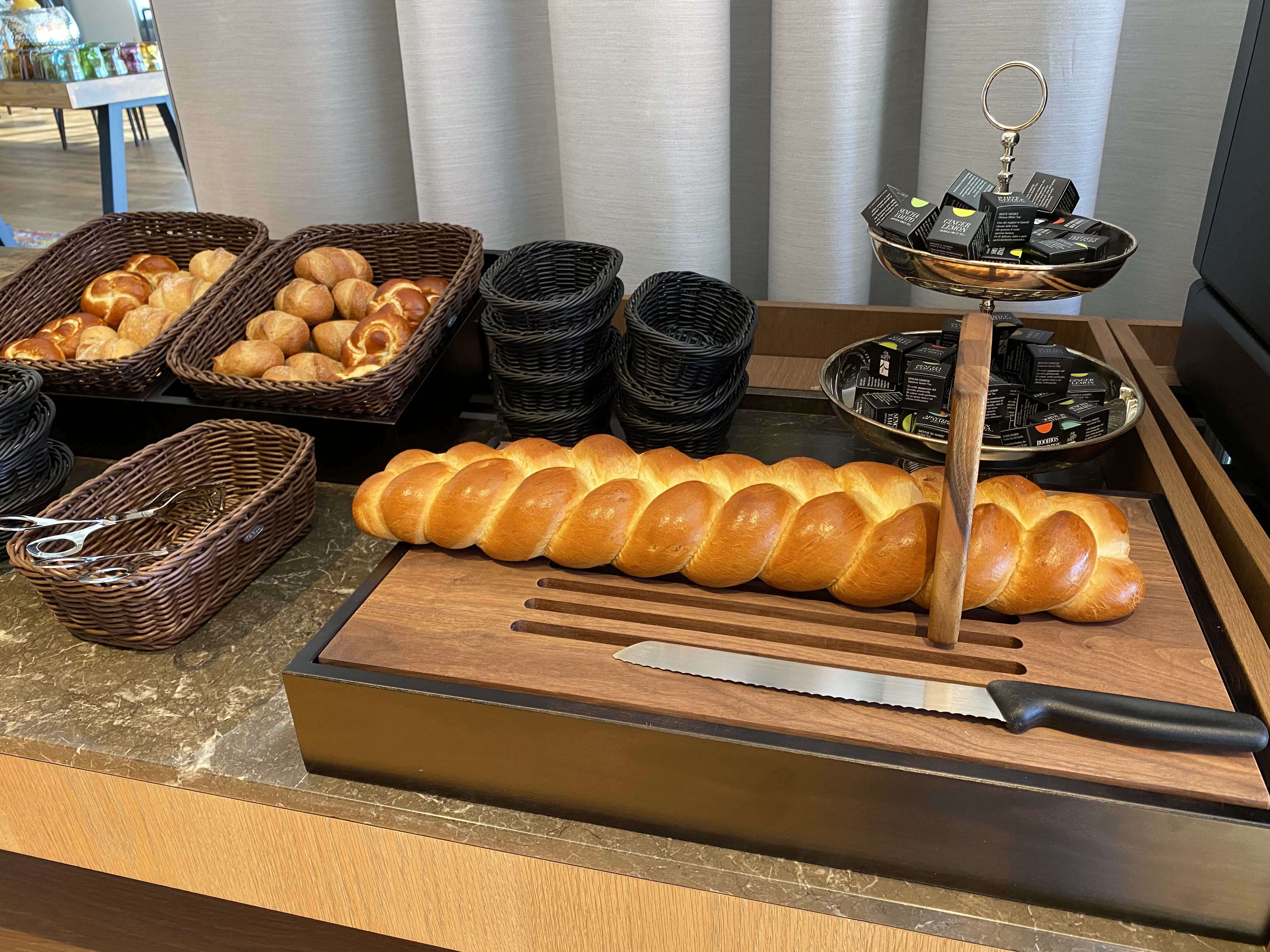 Buffet-Szene eines Macardo Brunchs: Körbe mit Brötchen, geflochtener Zopf auf Holzbrett mit Messer, Teeboxen auf Etageren und Servierzange. Alles für einen gemütlichen Brunch.
