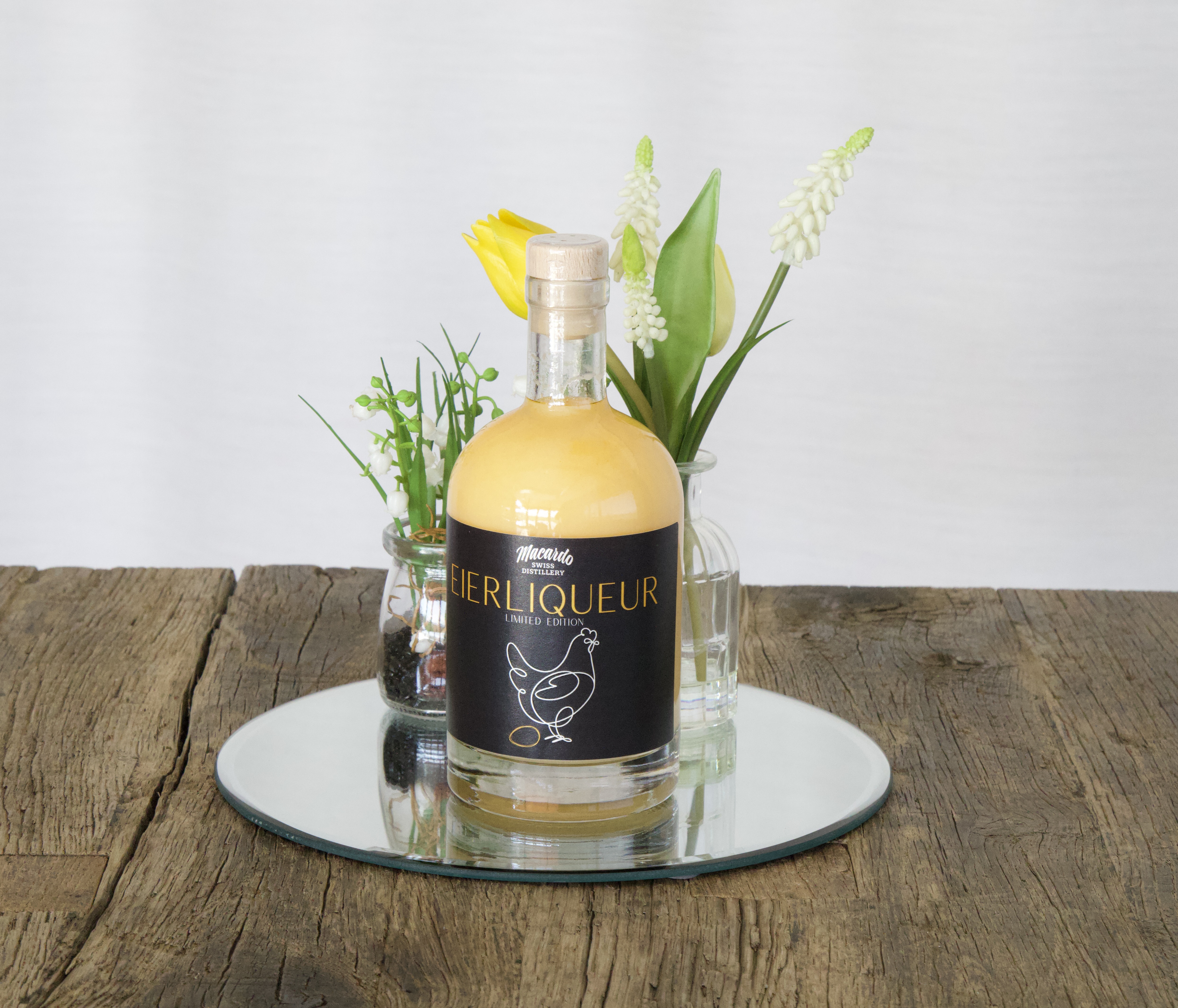Auf einem rustikalen Holztisch steht ein runder Spiegel. Darauf eine Flasche Eierlikör mit dunklem Etikett, das eine stilisierte Henne zeigt. Links ein kleines Glasgefäß mit Dekogrün, rechts eine Vase mit einer gelben Tulpe und weißen Blüten.