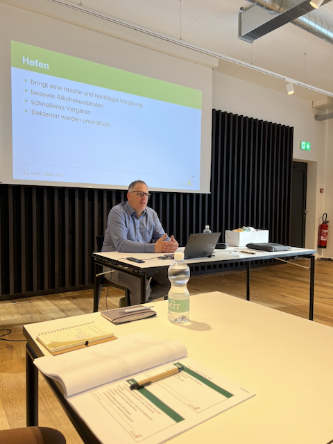 Innenaufnahme eines Raumes während des Macardo Brennkurses. Ein Mann sitzt an einem Tisch vor einer Leinwand mit "Hefen". Auf einem weiteren Tisch liegen Kursunterlagen. Das Licht ist hell. Der Raum wirkt modern.