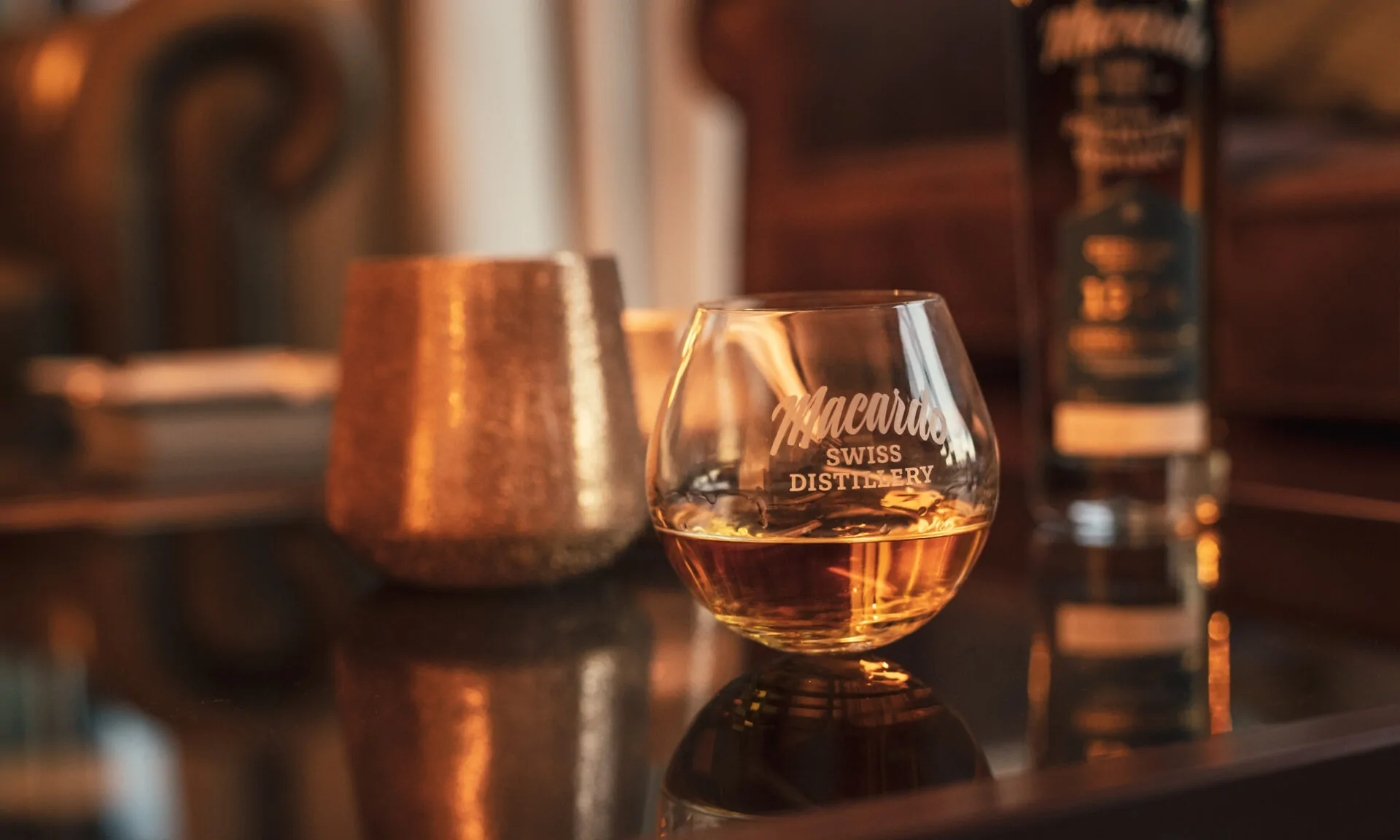 Nahaufnahme eines Whiskyglases mit der Aufschrift "Macardo Swiss Distillery", daneben eine goldfarbene Dose. Im Hintergrund eine unscharfe Whiskeyflasche und ein Ledersessel. Alles steht auf einer glänzenden Oberfläche.