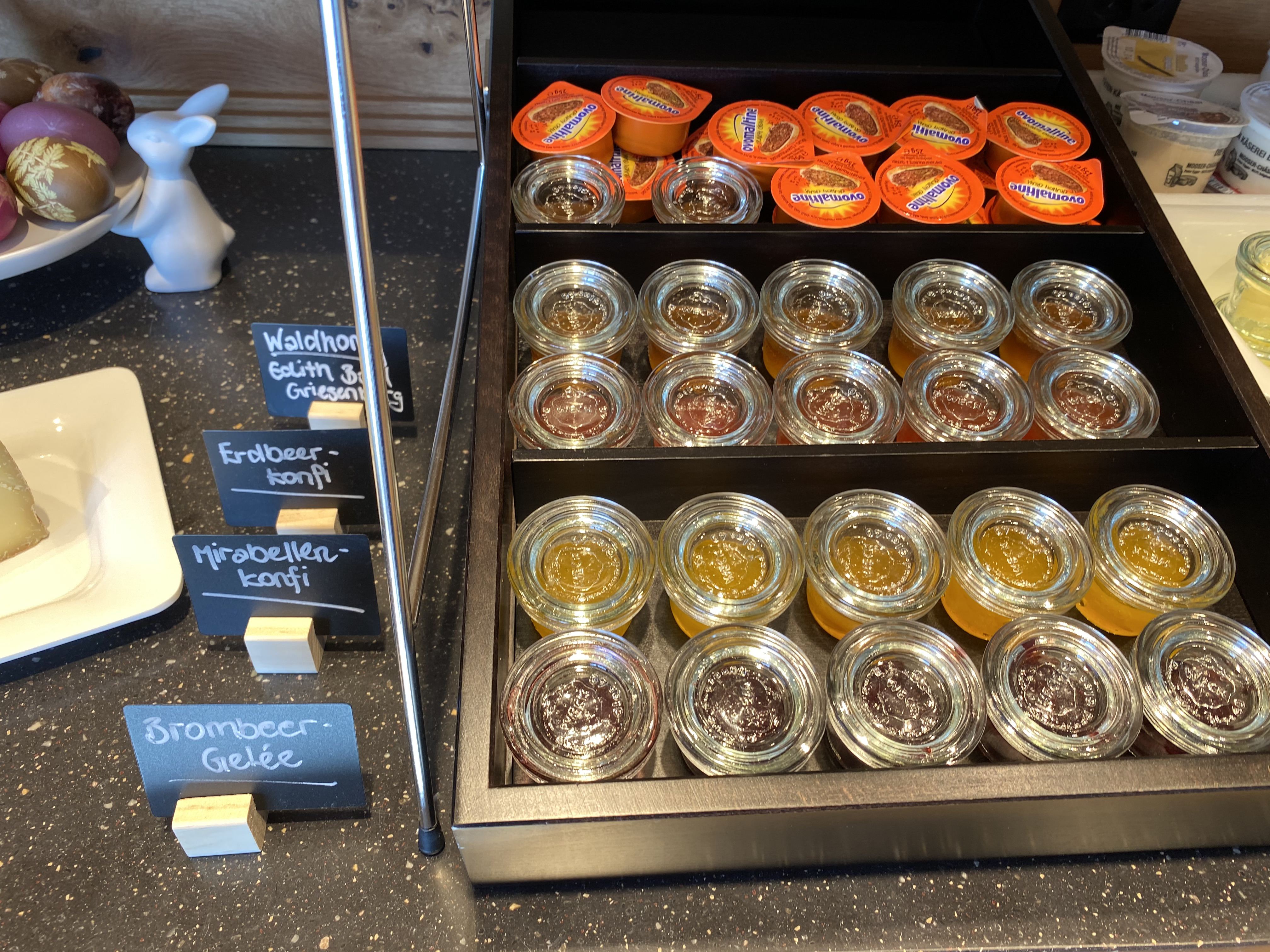 Auf dem Brunch-Buffet: Eine Etagere mit Ovomaltine Creme, diversen Konfitüren und Gelees wie Erdbeer, Mirabellen, Brombeer. Daneben ein Schild für Waldhonig, ein Keramik-Hase und ein Teller mit bemalten Eiern.