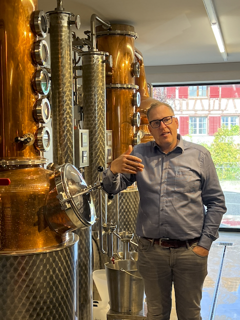 Ein Mann mit Brille und blauem Hemd steht vor kupferfarbenen Destillierkolben in einer Macardo Swiss Distillery. Er gestikuliert und erklärt den Brenn-Prozess während eines Brennkurses. Im Hintergrund ist ein Fenster mit Blick auf Gebäude zu sehen.