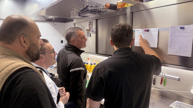 In einer Küche stehen vier Männer und betrachten eine Liste, die an einer Edelstahltür hängt. Ein Mann in Kochkleidung deutet mit dem Finger auf die Liste. Die Liste scheint Zutaten für ein Rezept zu enthalten. Eine weitere Liste hängt daneben.