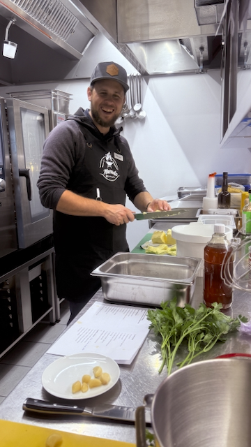 In einer Küche steht ein lächelnder Koch, der Butter schneidet. Auf der Arbeitsfläche liegen Petersilie, Knoblauchzehen und ein Rezept. Im Hintergrund sind Küchengeräte zu sehen. Fokus: Zubereitung beim Kochkurs.