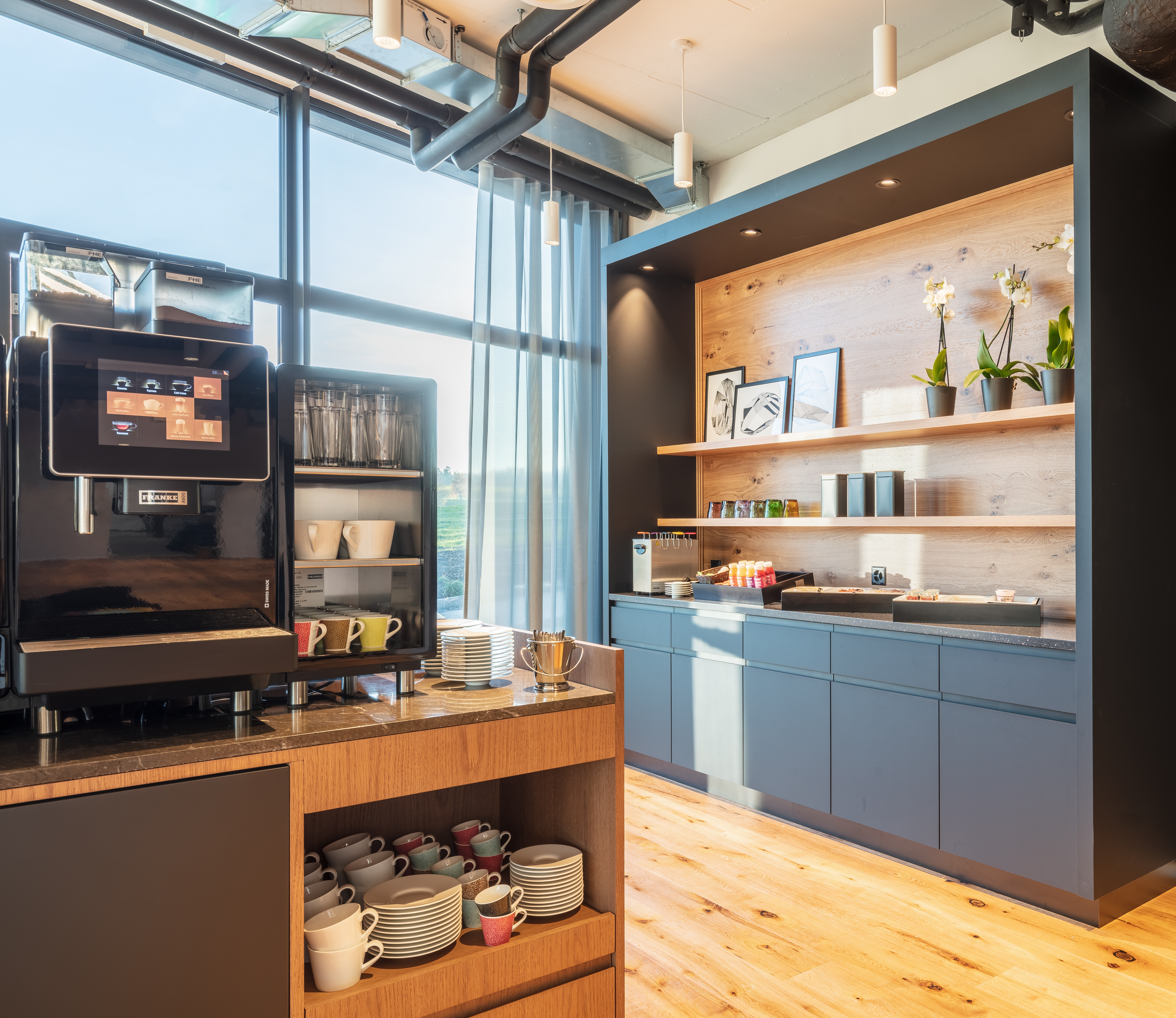 Innenansicht vom Buffetbereich des Macardo Brunch, mit Kaffeevollautomat, Gläser, Tassen, Tellern und einer Auswahl an Speisen auf einem Regal mit Holzverkleidung. Sonnenlicht durchflutet den Raum durch ein großes Fenster.