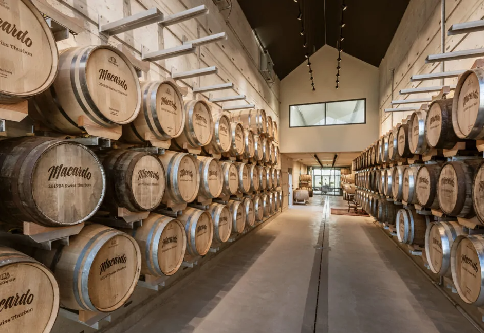 Innenaufnahme eines Lagerraums mit hellgrauen Betonwänden. Holzfässer mit Aufschrift "Macardo" sind in Reihen auf Metallregalen an den Wänden gestapelt. Ein Gang führt zu einem Fenster am Ende. Dunkelgraue Decke mit Hängelampen.