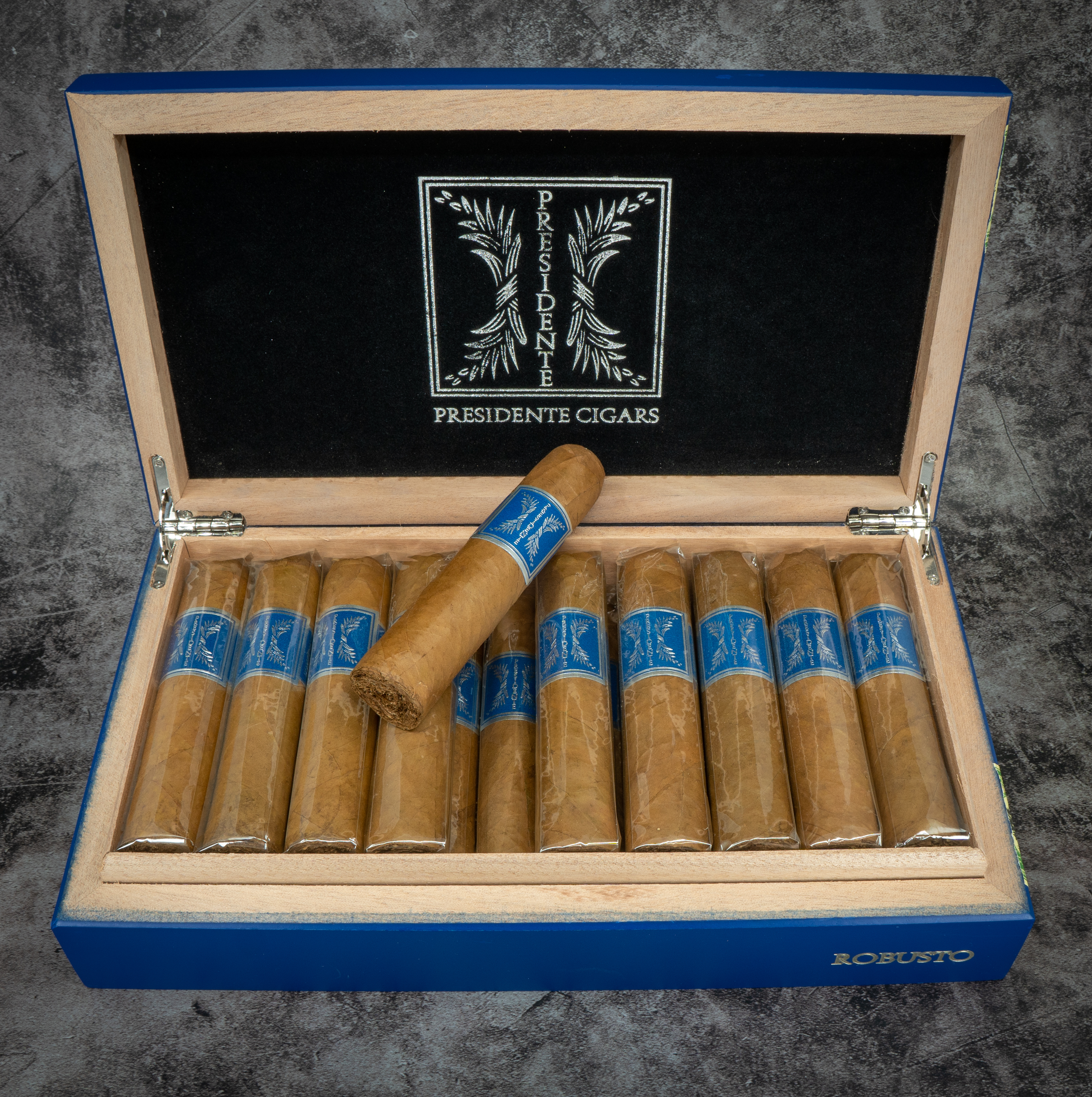 Geöffnete blau lackierte Holzkiste mit hellen Zigarren, einzeln in Folie verpackt. Eine Zigarre liegt quer darüber. Deckel mit schwarzem Innenfutter und silbernem Schriftzug „Presidente Cigars“. Auf der Kiste steht „Robusto“.